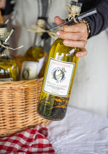 Load image into Gallery viewer, Person holding a bottle of artisan olive oil with a label, surrounded by other bottles and a basket.