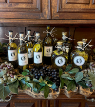 Load image into Gallery viewer, Bottles of olive oil with labels on a wooden surface with grapes and leaves.
